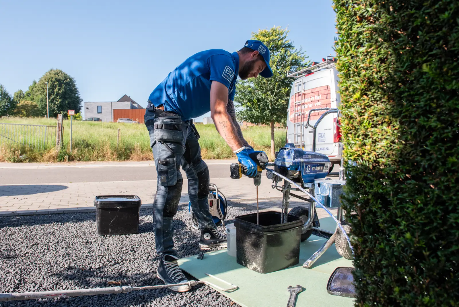 Een man in blauwe werkkleding en handschoenen mengt buiten, naast een bestelbusje en apparatuur, een stof in een zwarte bak, met groen en gebouwen op de achtergrond.