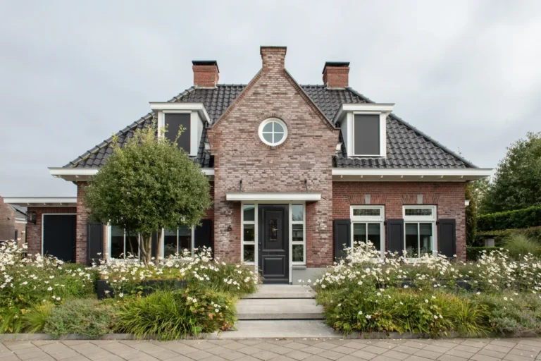 Een bakstenen huis met zwarte dakpannen, ramen met witte kozijnen, een zwarte voordeur, twee dakkapellen en een tuin met witte bloemen en groene struiken aan de voorkant.