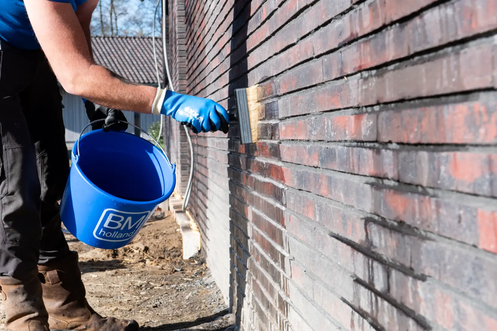 Een persoon met blauwe handschoenen aan is bezig met een kwast op een bakstenen muur, terwijl hij een blauwe BM Holland-emmer vasthoudt. De scène speelt zich buiten af; een deel van zijn arm en zijn laarzen zijn te zien.
