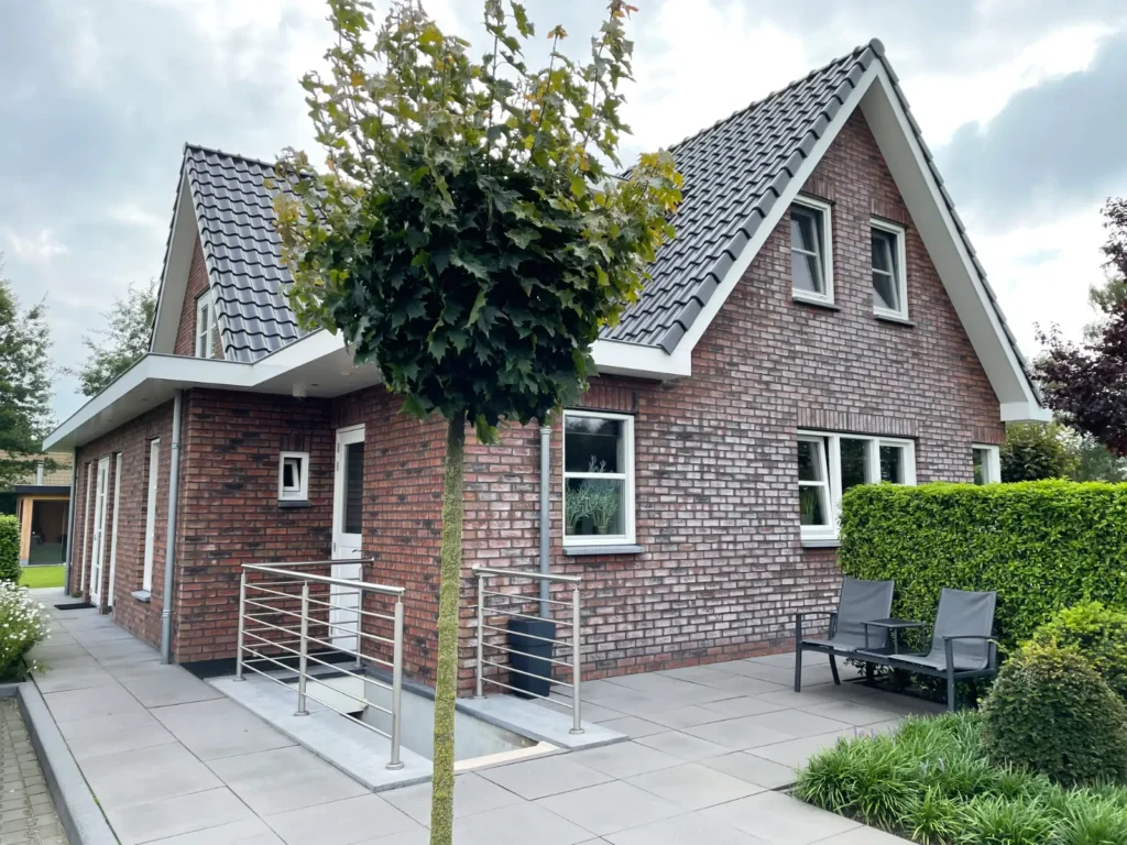 Een moderne bakstenen woning met een schuin grijs dak, metalen leuningen en grote ramen. Op het betegelde terras staan twee zwarte stoelen, er is een keurig gesnoeide haag en op de voorgrond staat een loofrijke boom.