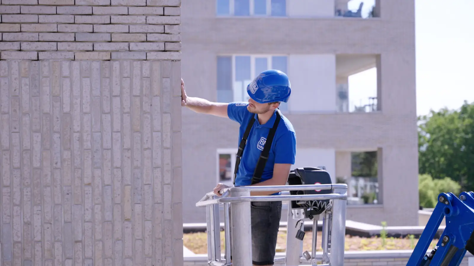 Een bouwvakker met een blauwe helm en een blauw shirt inspecteert een bakstenen muur terwijl hij op een verhoogd platform staat; op de achtergrond is een modern gebouw te zien.