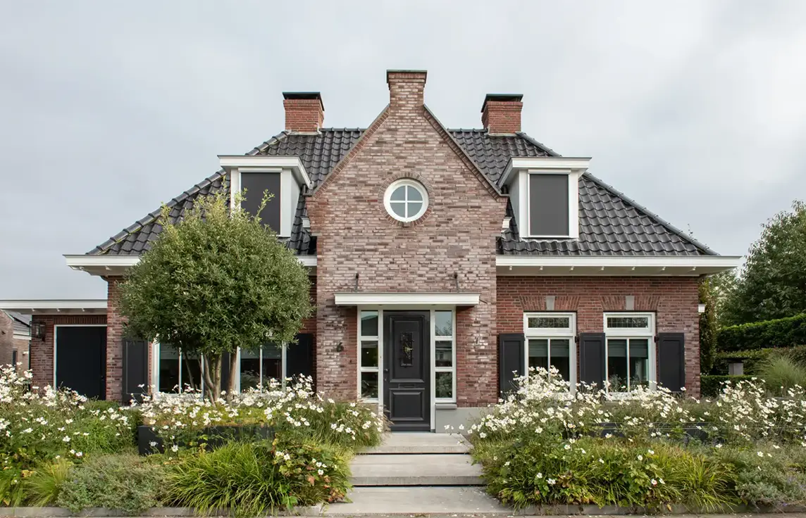 Een bakstenen huis van twee verdiepingen met zwarte dakpannen, ramen met witte kozijnen, zwarte luiken, een centraal geplaatste voordeur en een tuin met witte bloemen en groene struiken aan de voorkant.