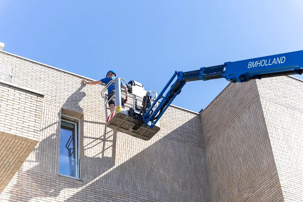 Een werknemer in een blauw overhemd en met een helm op gebruikt een blauwe BM HOLLAND-lift om onder een strakblauwe hemel aan de buitenmuur van een lichtgekleurd bakstenen gebouw te werken.