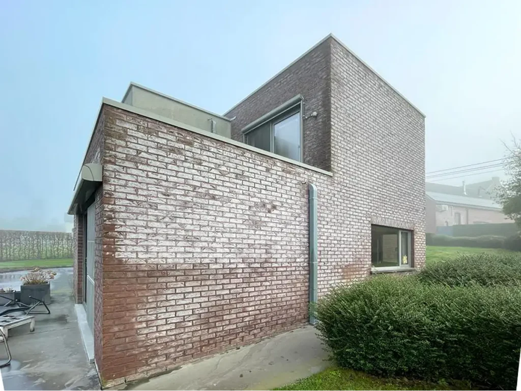 Modern bakstenen huis met grote ramen en een plat dak, gelegen op een betonnen terras met zitgelegenheid buiten. Het gebouw wordt omringd door struiken en gras; op de achtergrond is een mistige lucht te zien.
