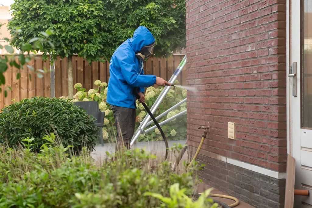 Iemand in een blauwe regenjas maakt met een hogedrukreiniger de bakstenen muur van een huis schoon, in een tuin met groene planten en een houten schutting op de achtergrond.
