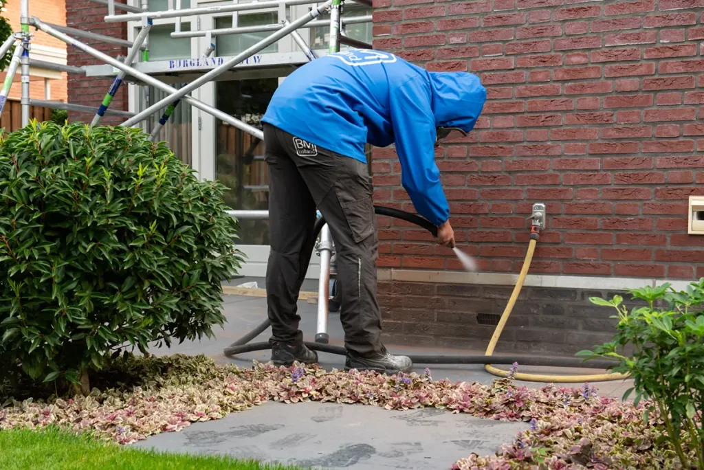 Een persoon in een blauw jasje maakt met een hogedrukreiniger een terras schoon naast een bakstenen gebouw, met groen en planten op de voorgrond.