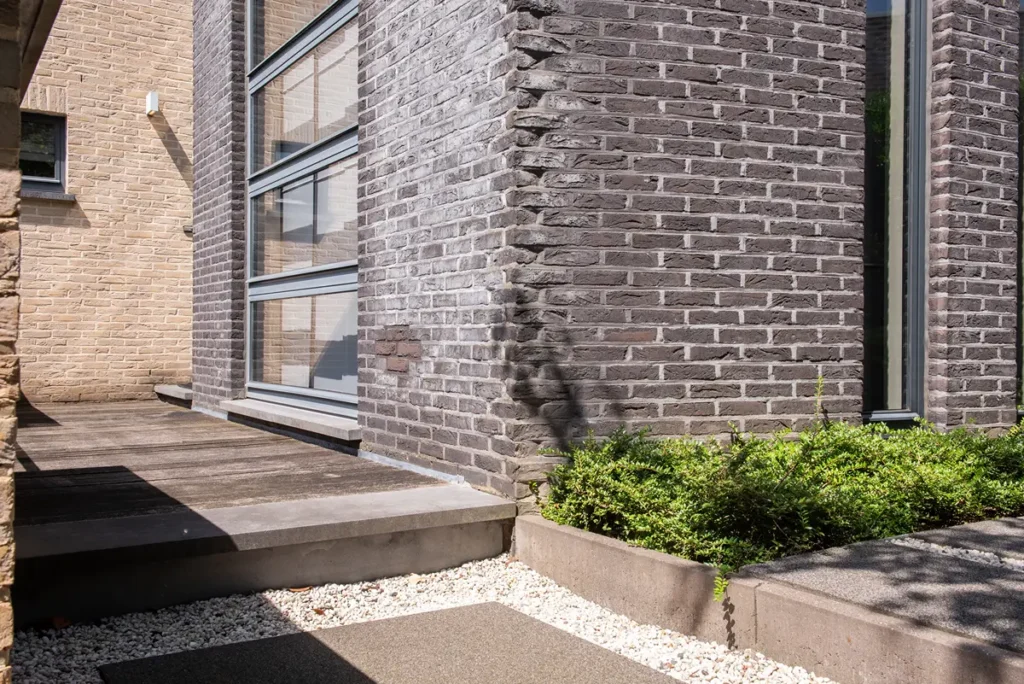 De buitenkant van een modern gebouw met grijze bakstenen muren, grote ramen, een houten pad, betonnen treden, wit grind en groene struiken in een verhoogde plantenbak, badend in fel zonlicht.