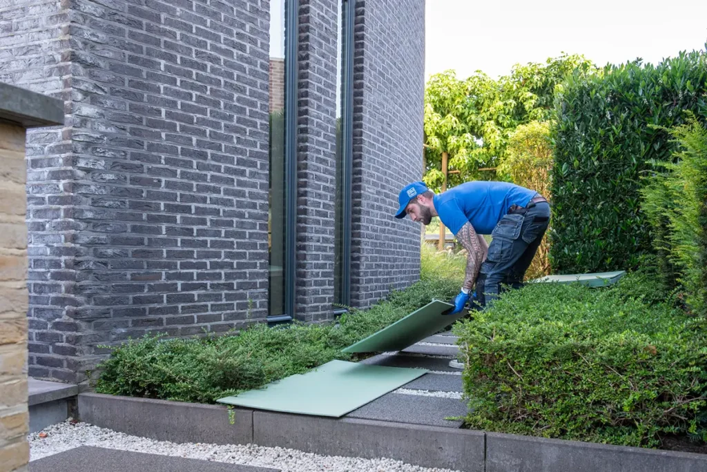 Iemand in een blauw shirt en een pet legt grote groene planken over de struiken naast een modern bakstenen gebouw. De planken vormen een pad over de planten in een tuingedeelte.