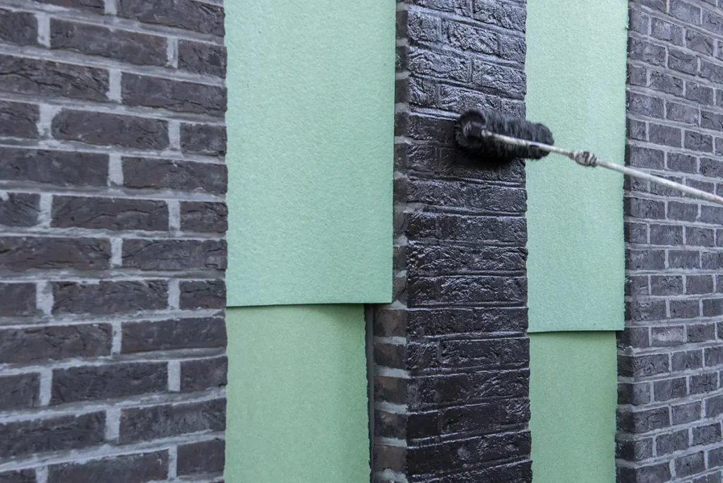Iemand brengt met een verfroller zwarte verf aan op een bakstenen muur. Groene rechthoekige vellen papier bedekken delen van de muur, waarschijnlijk om deze tegen verf te beschermen. Het tafereel doet denken aan een schilder- of renovatieproject.