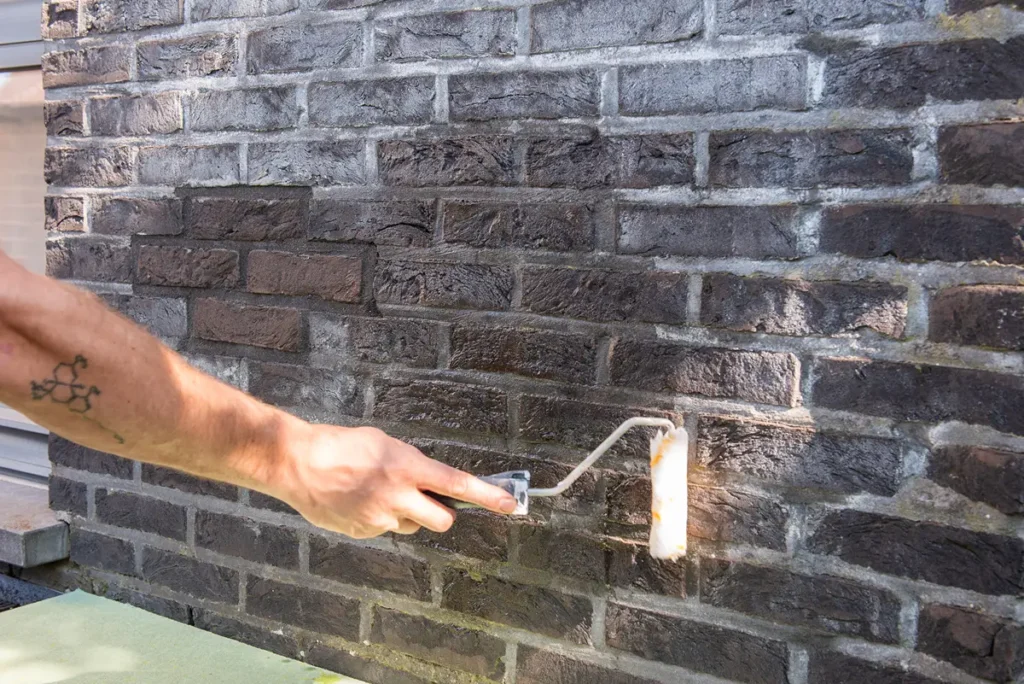 Iemand brengt met een verfroller een heldere vloeistof aan op een donkere bakstenen muur, waarschijnlijk om het oppervlak af te dichten of waterdicht te maken.