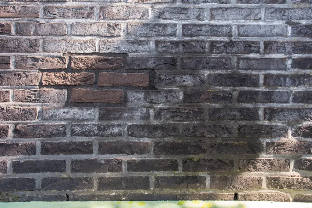 Een bakstenen muur met overwegend donkerbruine stenen, die ongelijkmatig van kleur zijn, met hier en daar een paar lichtere en meer roodachtige stenen linksboven. Zonlicht en schaduwen zorgen voor lichte en donkere plekken over het hele oppervlak.