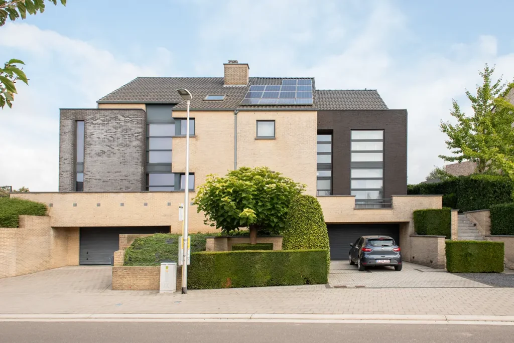 Moderne woning met twee verdiepingen, opgetrokken uit baksteen met donkergrijze accenten, grote ramen, zonnepanelen op het dak, een auto die op de oprit geparkeerd staat en keurig gesnoeide struiken en bomen aan de voorkant.