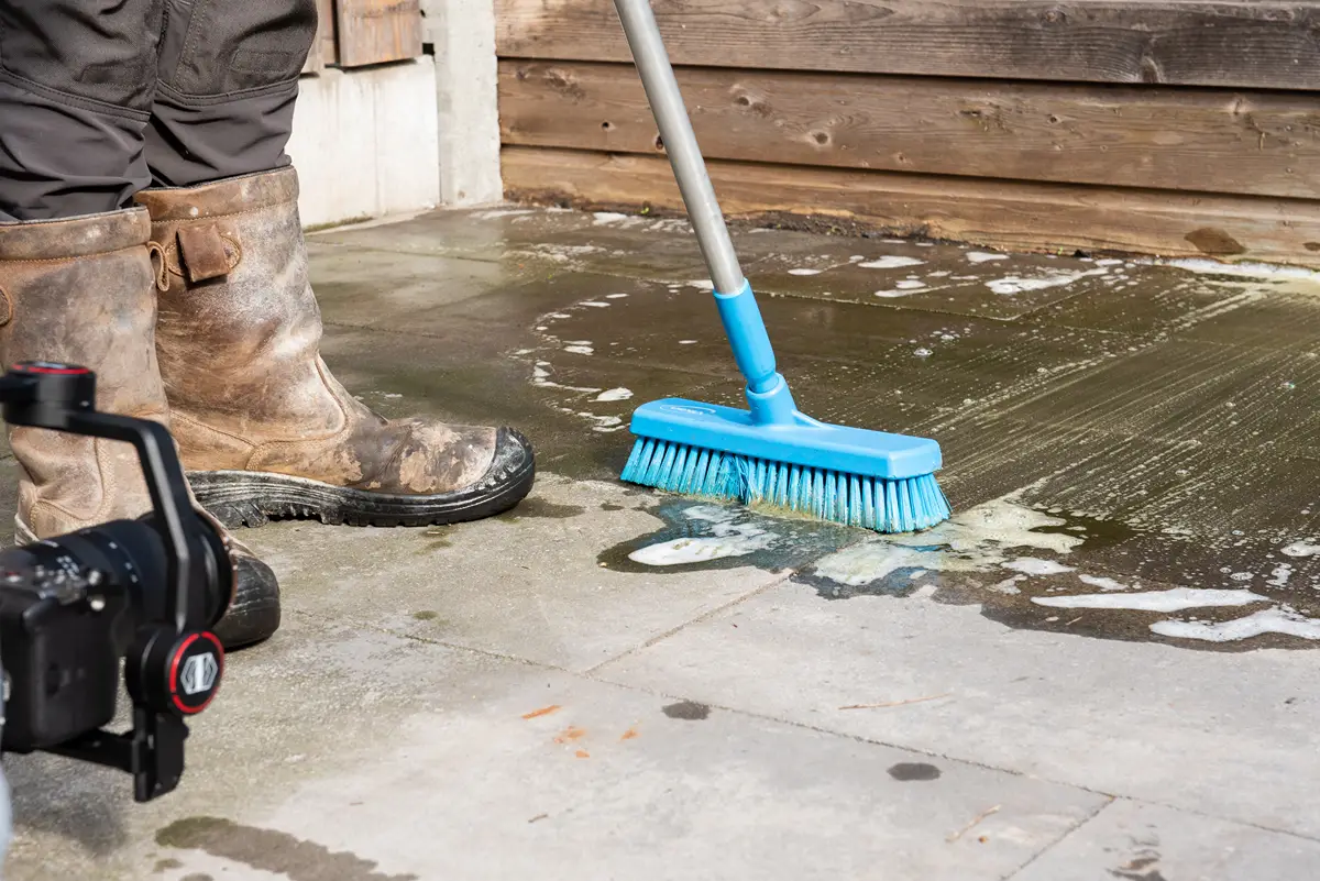 Iemand met modderige laarzen schrobt een met zeep ingesmeerd terras schoon met een blauwe borstel aan een steel. In de linkeronderhoek is een camera te zien die het schoonmaakproces vastlegt.