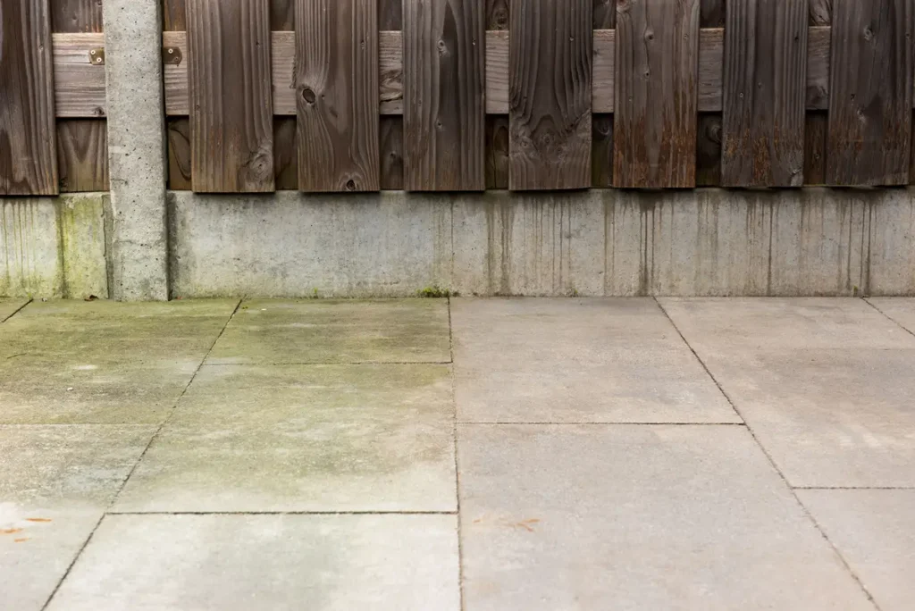 Betonnen terrastegels met hier en daar wat mos en verkleuringen aan de linkerkant, met op de achtergrond een houten schutting die op een betonnen fundering staat.