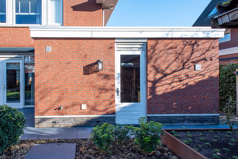 Een witte deur in een rode bakstenen muur van een moderne woning, met een raam en een buitenlamp ernaast. De schaduwen van boomtakken vallen over de muur en op de voorgrond zijn bloembedden te zien.