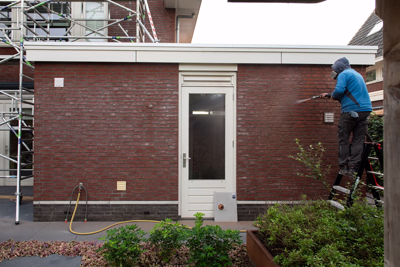 Een persoon in een blauw jasje reinigt met een hogedrukreiniger een rode bakstenen muur naast een witte deur. Links staat een steiger en op de voorgrond staan planten.