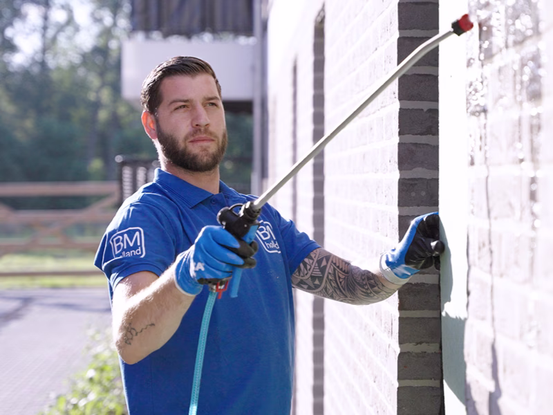 Een man in een blauw overhemd en handschoenen spuit buiten vloeistof op een bakstenen muur; hij houdt een lange spuitstang vast en is in het daglicht aan het werk.