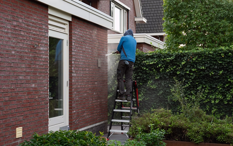 Een persoon in een blauwe hoodie staat op een ladder en maakt met een hogedrukreiniger de bakstenen buitenmuur van een huis schoon, vlakbij een groene haag en een tuin.