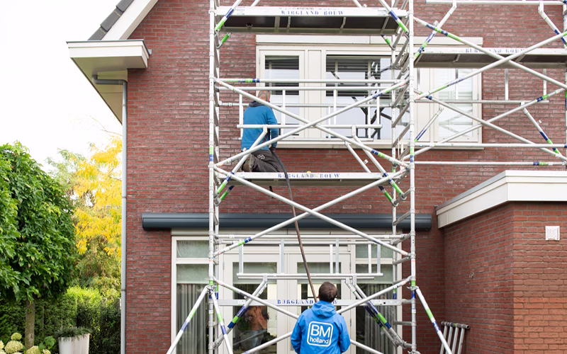 Twee arbeiders staan op een steiger buiten een bakstenen huis en voeren onderhouds- of bouwwerkzaamheden uit bij een raam op de bovenverdieping. De steiger beslaat het grootste deel van de voorgevel van het huis. Aan de linkerkant is gedeeltelijk een boom te zien.