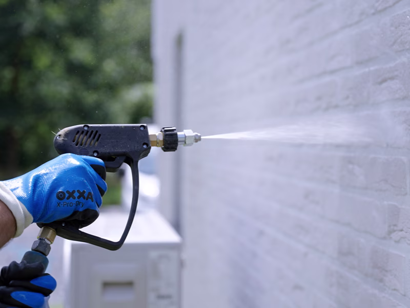 Iemand met blauwe handschoenen aan maakt een witte bakstenen muur schoon met een hogedrukreiniger, waarbij hij water in een gerichte straal spuit.