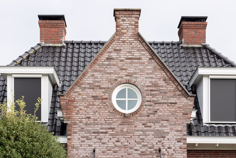 Een close-up van een bakstenen huis met een rond raam in het midden, twee schoorstenen, een zwart pannendak en twee dakkapellen met gesloten luiken aan weerszijden.