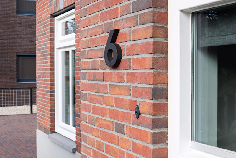 De buitenkant van een bakstenen huis met een groot zwart cijfer 6 aan de muur naast een raam met een wit kozijn.