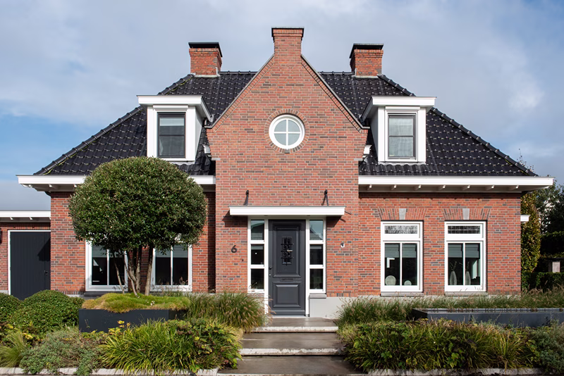 Een huis van rode baksteen met een zwart pannendak, dakkapellen, een rond raam in het midden, een grijze voordeur, twee schoorstenen en mooi aangelegde struiken en planten in de voortuin.