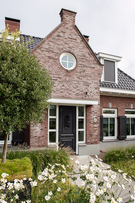 Een bakstenen huis met een puntdak, een rond raam boven de deur, zwarte luiken en een tuin met witte bloemen op de voorgrond.