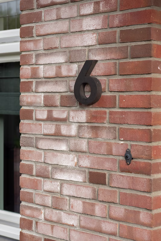 Op een rode bakstenen muur naast een raam is een zwart cijfer 6 aangebracht.