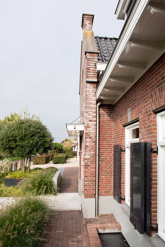 Een bakstenen huis met zwarte luiken en een pannendak staat naast een aangelegd voetpad, met groene struiken en bomen, en een heldere hemel op de achtergrond.