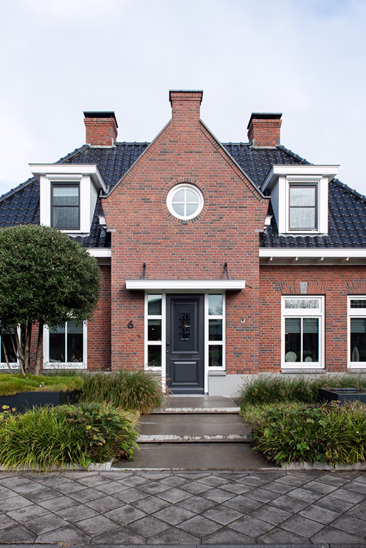Een huis van rode baksteen met een grijze deur in het midden, een rond raam erboven, een zwart pannendak, twee schoorstenen, ramen met witte kozijnen, groene struiken en een betegeld pad dat naar de ingang leidt.