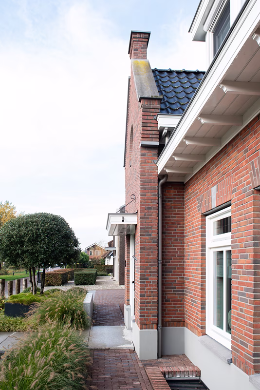 Een bakstenen huis met wit omrande ramen en een donker betegeld dak, omgeven door groen en een bakstenen pad, onder een gedeeltelijk bewolkte hemel.