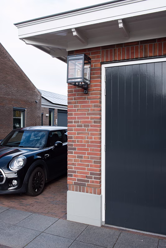 Er staat een zwarte auto geparkeerd op een bakstenen oprit naast een bakstenen gebouw met een donkergrijze deur en een buitenwandlantaarn.