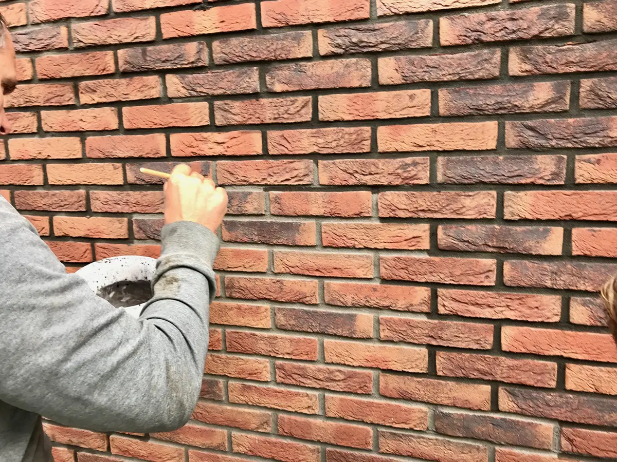 Een persoon in een grijze trui brengt met een klein penseel materiaal aan op de mortelvoegen van een rode bakstenen muur; waarschijnlijk is hij bezig met reparatie- of onderhoudswerkzaamheden.