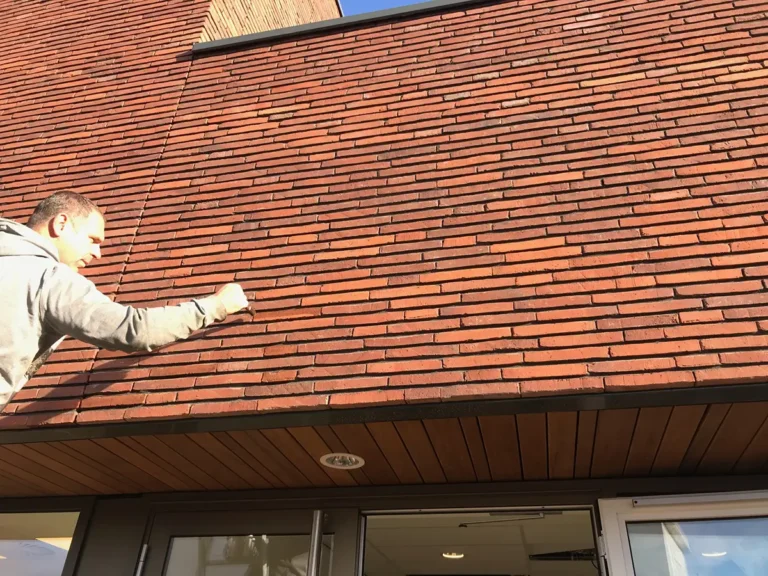 Links staat iemand die lijkt te schilderen of een schuine bakstenen muur aanraakt, terwijl het zonlicht schaduwen werpt en eronder een houten overkapping met lampen te zien is.