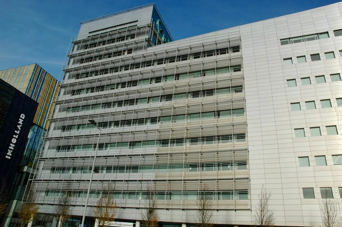 Een hoog, modern kantoorgebouw met meerdere verdiepingen, grote ramen en horizontale metalen lamellen, afgetekend tegen een helderblauwe lucht. Voor het gebouw zijn hier en daar wat bomen en een lantaarnpaal te zien.