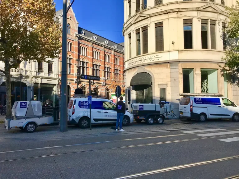 Drie witte bestelwagens van BM Solutions met aanhangwagens staan geparkeerd in een straat in de stad, vlakbij een gebogen gebouw. Tussen de voertuigen staat iemand met een witte helm en een blauw vest. Op de achtergrond zijn bomen en gebouwen in oude stijl te zien.