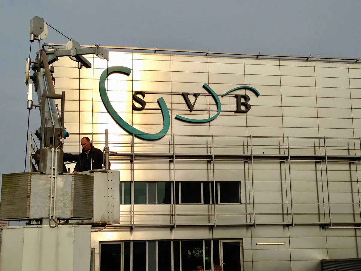 Twee werknemers op een hoogwerkplatform voeren onderhoudswerkzaamheden uit bij de buitenmuur van een modern gebouw, waarop grote „SVB“-letters en een blauwgroen swoosh-logo te zien zijn. Het gebouw is voorzien van reflecterende metalen panelen en ramen.