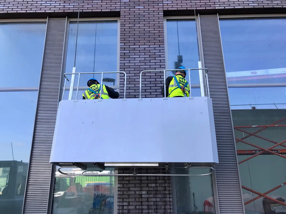 Twee bouwvakkers in veiligheidsvesten en helmen staan op een hangend platform voor een modern bakstenen gebouw met grote glazen ramen.