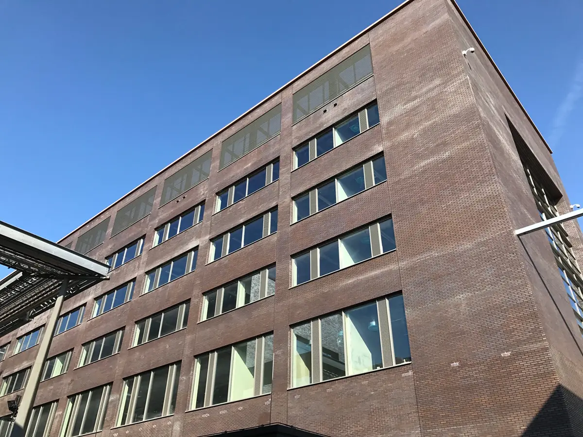 Een bruin bakstenen gebouw met grote ramen en een plat dak is te zien tegen een helderblauwe lucht. Het perspectief is van onderaf, waarbij men omhoog kijkt naar de hoek van het gebouw.