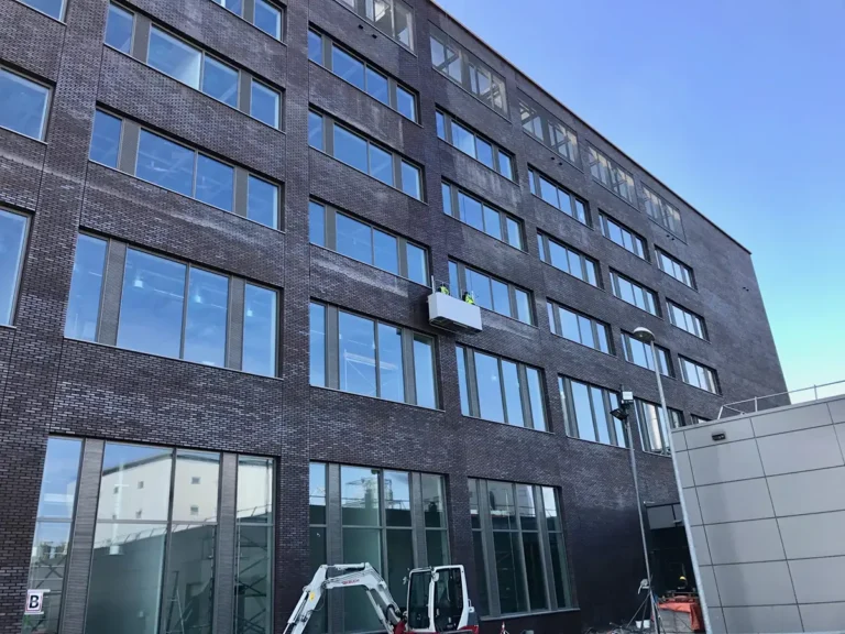 Een modern bakstenen gebouw met grote ramen, een bouwvoertuig dat buiten geparkeerd staat, en een balkon met twee mensen en planten dat uit een raam op een van de bovenste verdiepingen steekt.