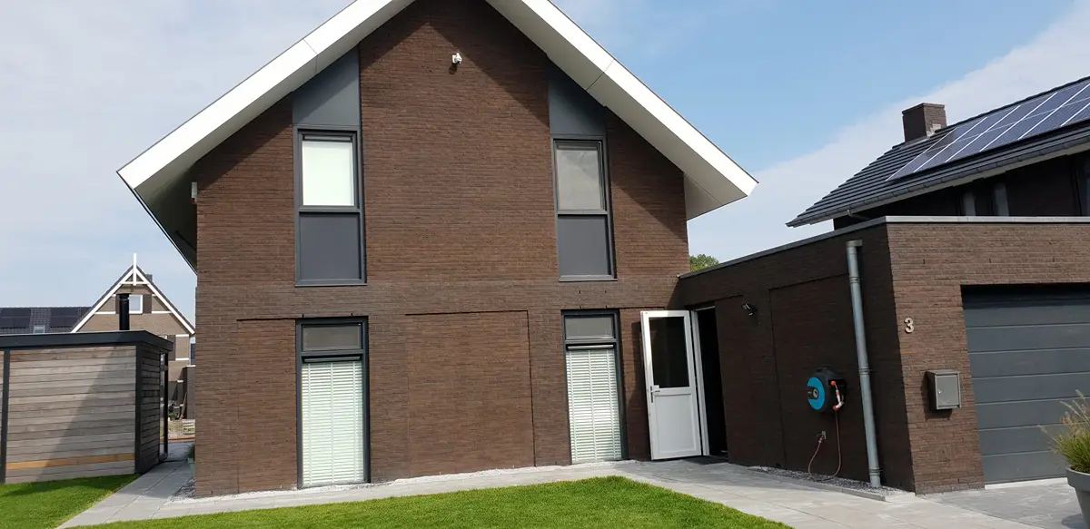 Een moderne bakstenen woning met twee verdiepingen, grijze raamkozijnen, witte sierlijsten en een kleine zij-ingang. De oprit en het gazon zijn zichtbaar, evenals een garage en zonnepanelen op het dak van een naburig huis.