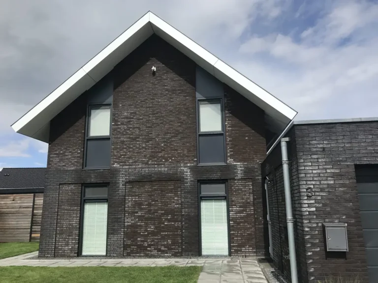 Moderne woning met twee verdiepingen, met een gevel van donkere baksteen, witte dakranden, vier verticale ramen en een betegeld voetpad dat naar de ingang leidt. De lucht is gedeeltelijk bewolkt.