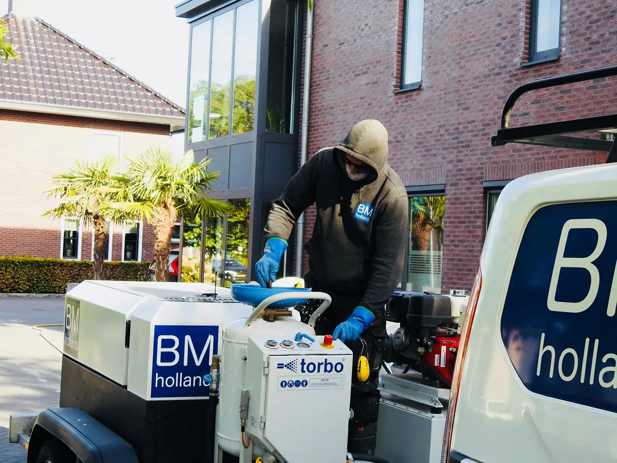Een werknemer in een hoodie en handschoenen bedient industriële reinigingsapparatuur buiten een bakstenen gebouw, met een servicebusje dat vlakbij geparkeerd staat.