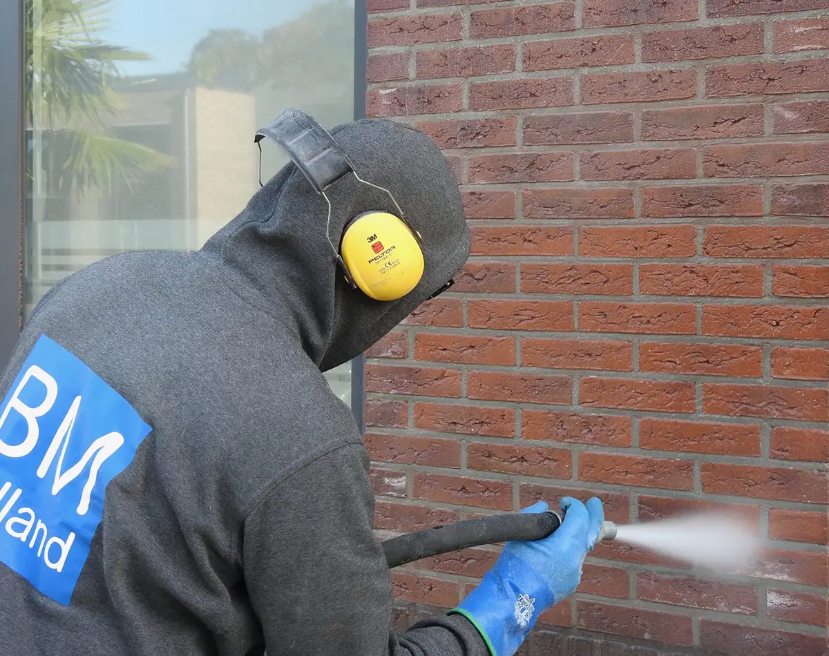 Iemand in een grijze hoodie, blauwe handschoenen en gele oorkappen is bezig een rode bakstenen muur naast een raam met een hogedrukreiniger schoon te spuiten.
