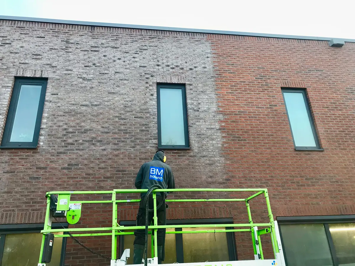 Een werknemer in een groene hoogwerker reinigt de gevel van een bakstenen gebouw, waardoor een felrood gedeelte zichtbaar wordt naast een nog steeds vuil, grijsachtig gedeelte. In het gedeelte dat wordt gereinigd, zijn twee hoge ramen te zien.