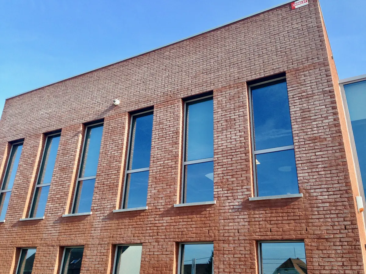 Een modern bakstenen gebouw met hoge rechthoekige ramen waarin de blauwe lucht weerspiegelt. De foto is vanuit een lage hoek genomen, waardoor de symmetrie van de ramen en de textuur van de bakstenen gevel goed tot hun recht komen.