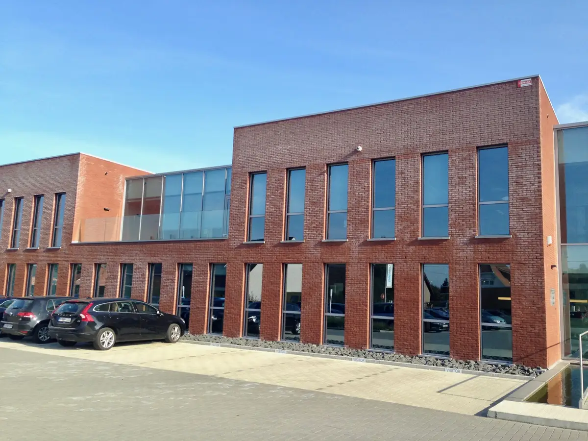 Een modern kantoorgebouw van baksteen met hoge ramen, een glazen gedeelte aan de linkerkant en geparkeerde auto’s ervoor op een zonnige dag.