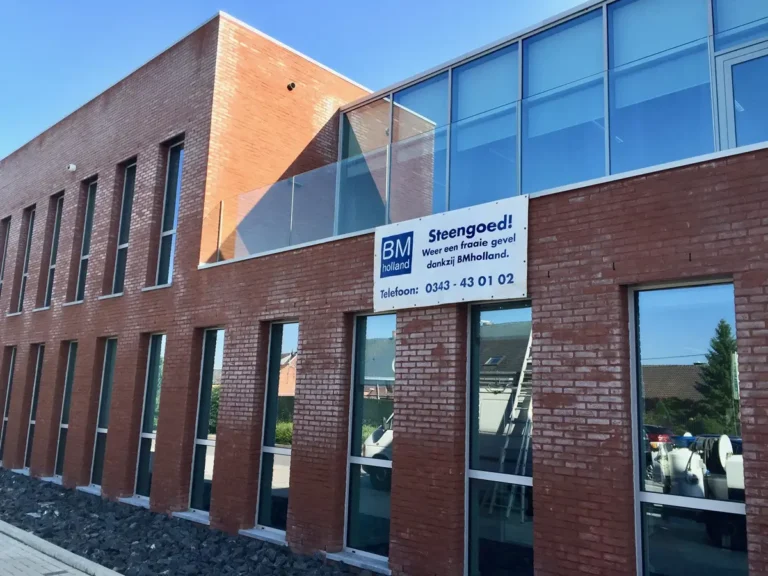 Een modern bakstenen gebouw met grote glazen ramen en een bord met de tekst „BM Holland Steengoed! Telefoon: 0343 - 43 01 02” op de gevel. Op de achtergrond zijn de blauwe lucht en de nabijgelegen huizen te zien.