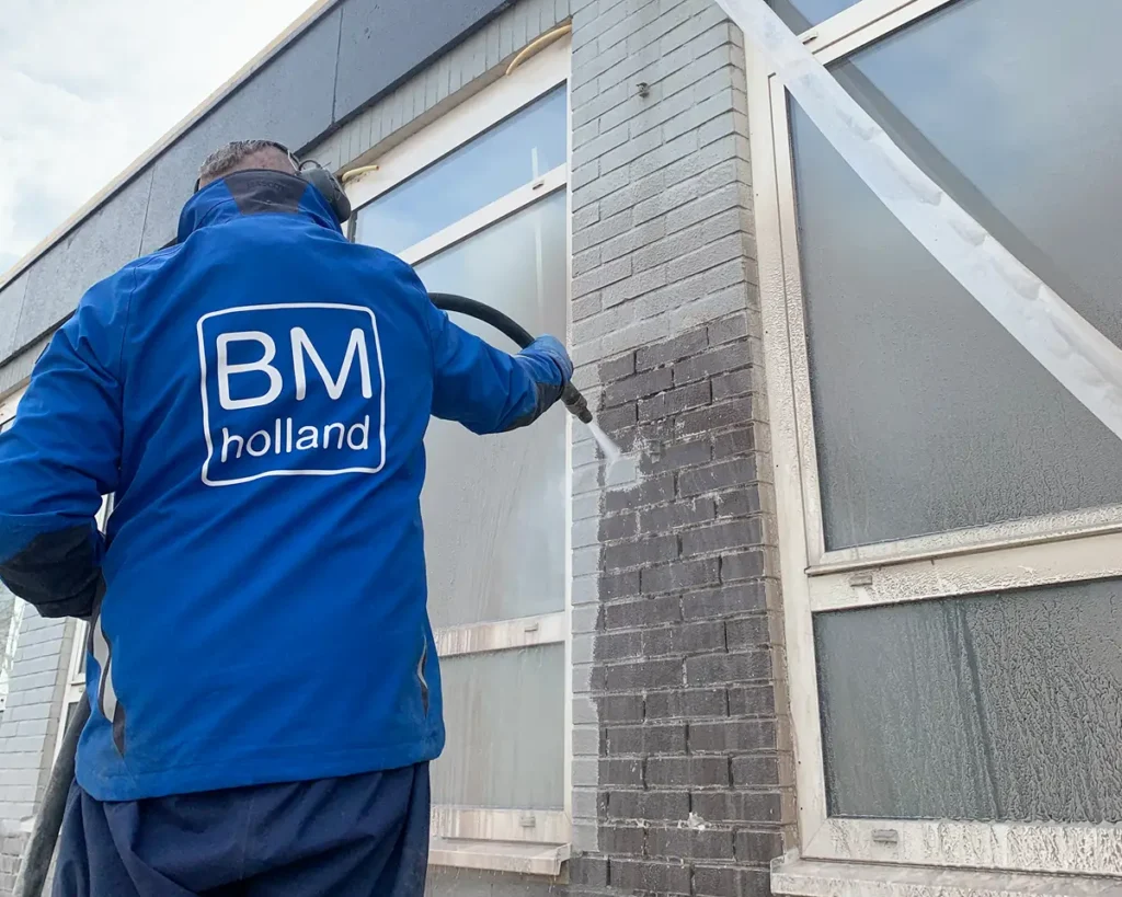 Een werknemer in een blauw BM Holland-jack maakt met een hogedrukslang de bakstenen aan de buitenkant van een gebouw schoon en verwijdert daarbij vuil of verf; naast het gereinigde gedeelte bevinden zich grote ramen.